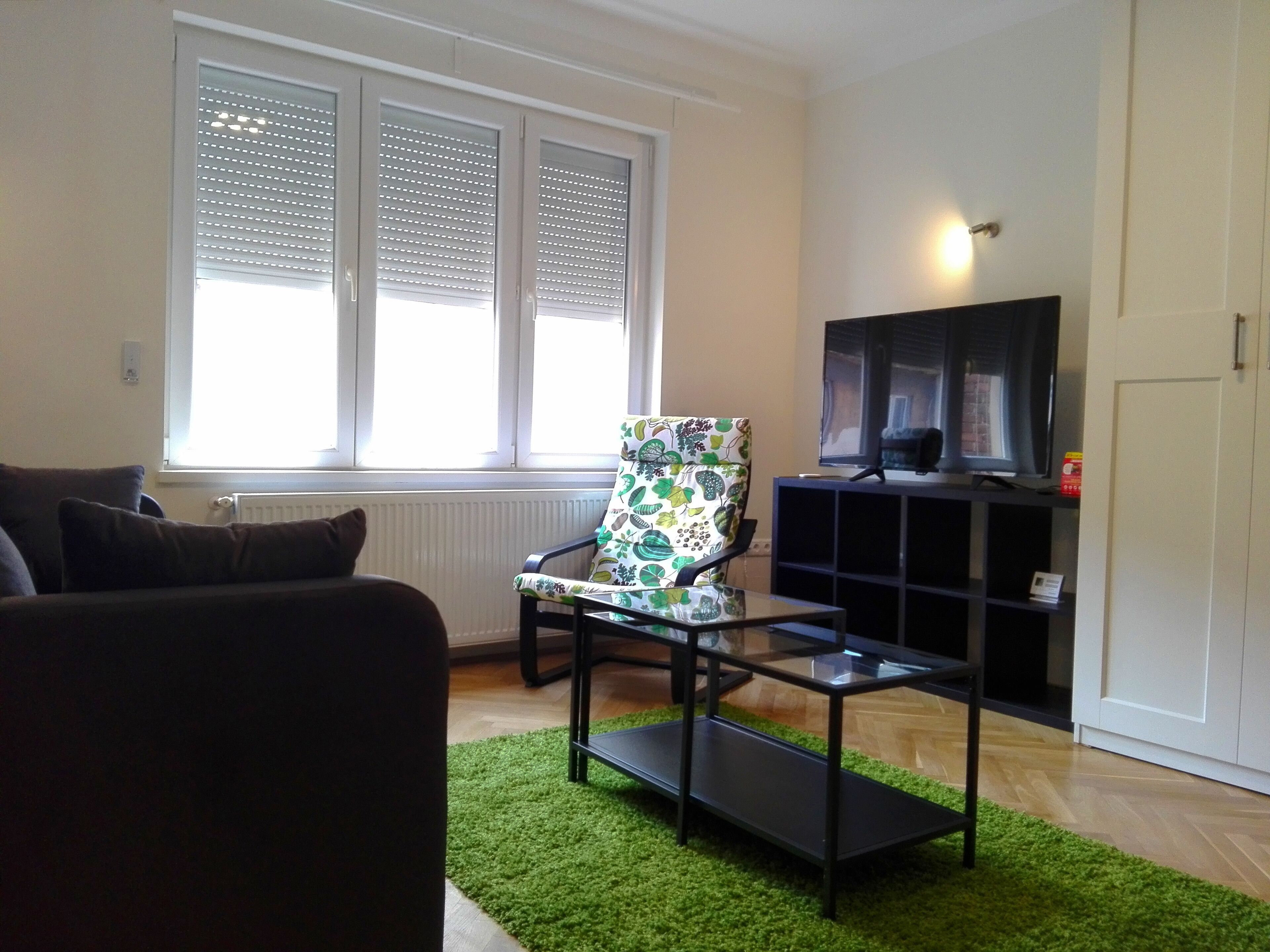 A living room with a sofa, a patterned armchair, a glass coffee table, a TV on a stand, and a radiator.