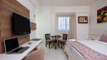 Habitación de hotel con cama, escritorio con computadora portátil, televisor, pequeña mesa con sillas y ventana con cortinas.