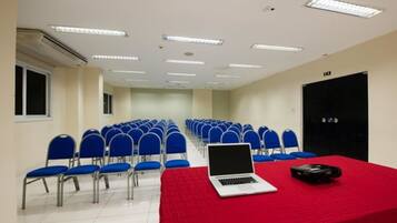 Un salón de conferencias con filas de sillas azules, una mesa roja con una computadora portátil y un proyector, y una puerta con un cartel.