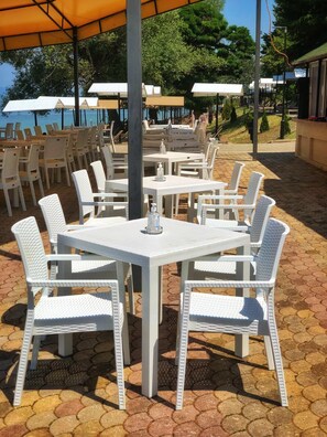 Terrasse mit weißen Tischen und Stühlen, Sonnenschirmen und einem befestigten Bereich.
