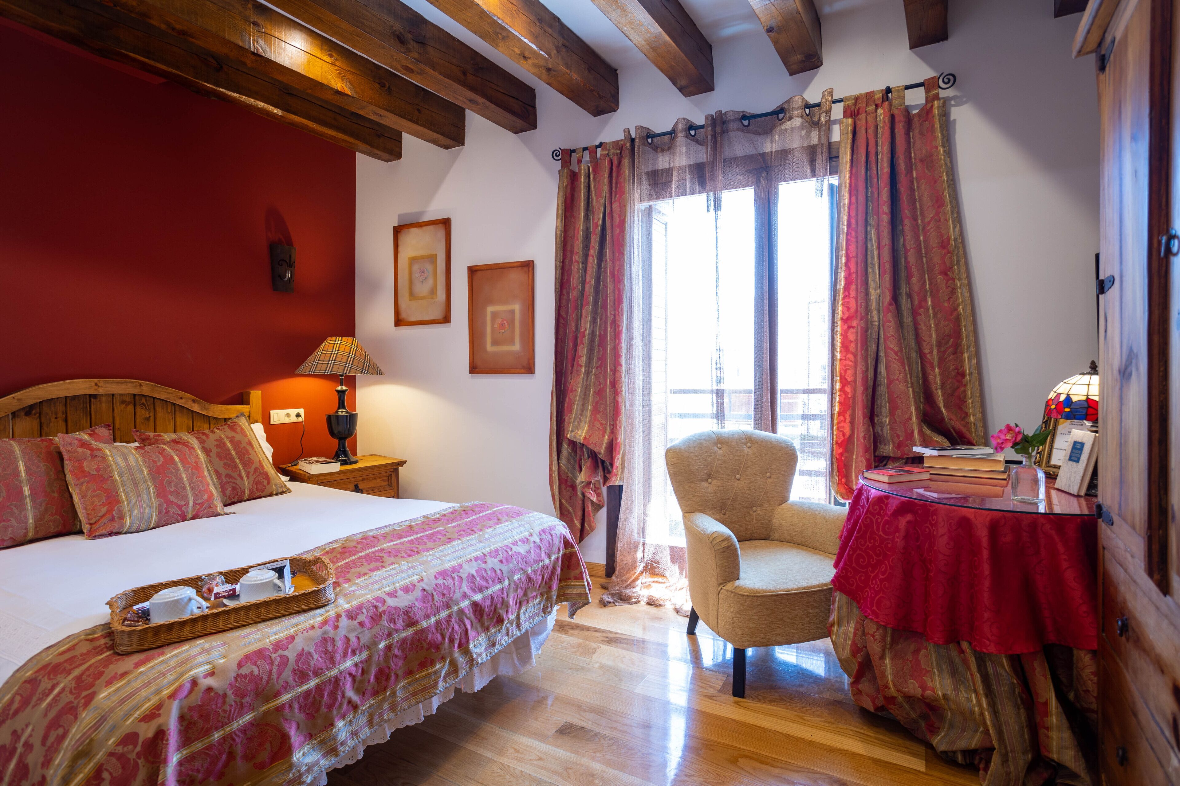 A bedroom with a wooden bed, a chair, a table with a red tablecloth, and a window with curtains.