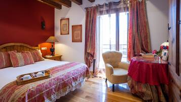 A bedroom with a wooden bed, a chair, a table with a red tablecloth, and a window with curtains.