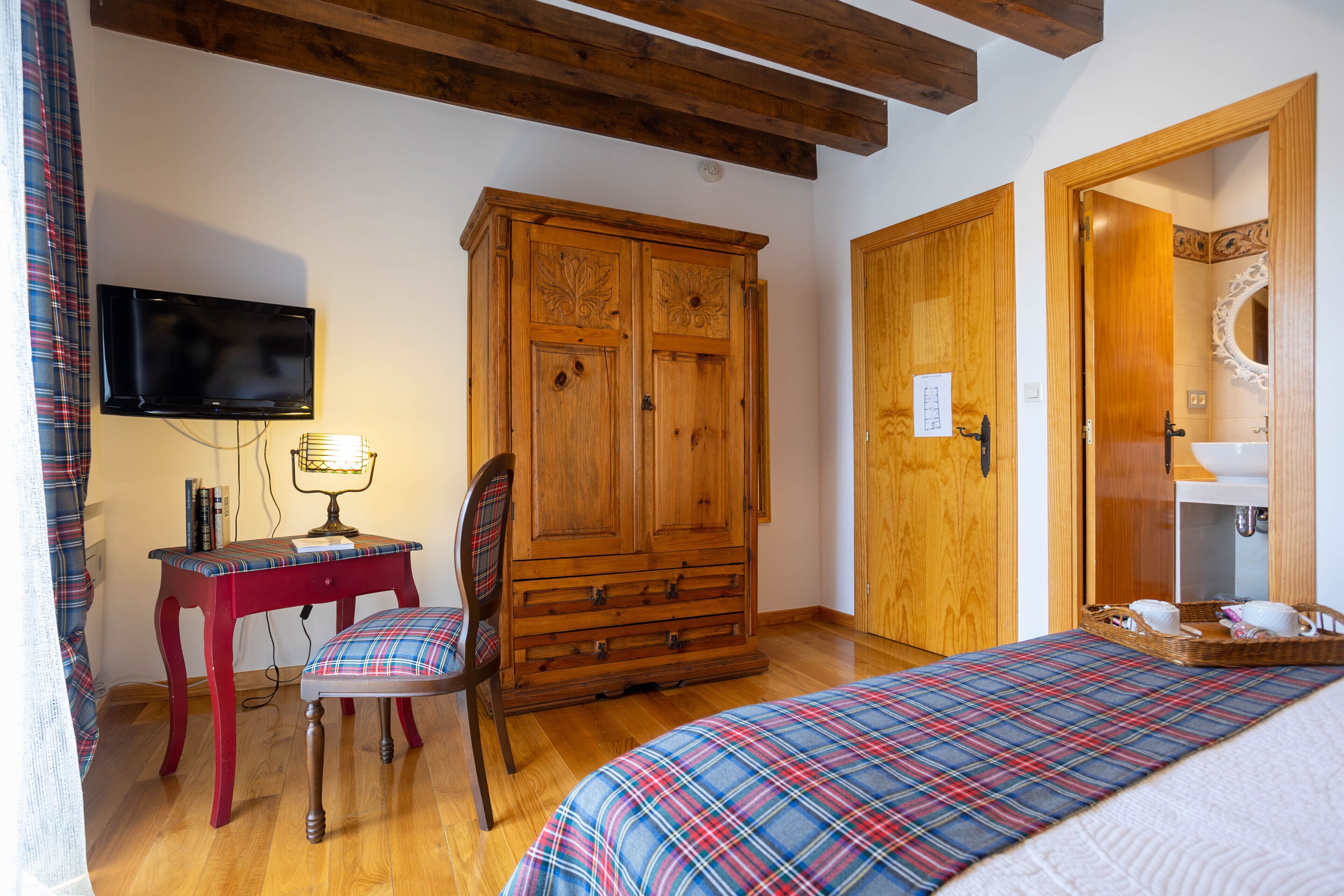 A bedroom with a wooden wardrobe, a television mounted on the wall, a desk with a chair, and a bed with a plaid bedspread.