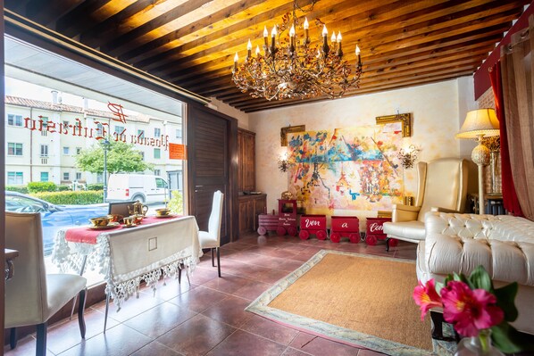 Quarto de hotel com teto de madeira, um candelabro, uma mesa de jantar com toalha vermelha, um sofá, uma pintura abstrata colorida e vista para a rua com um prédio e um carro.