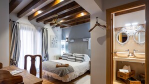 A bedroom with a bed, wooden ceiling beams, a ceiling fan, a bedside table, a nightstand, and a bathroom with a mirror, sink, and storage. - Posada De Eufrasio (Lerma)