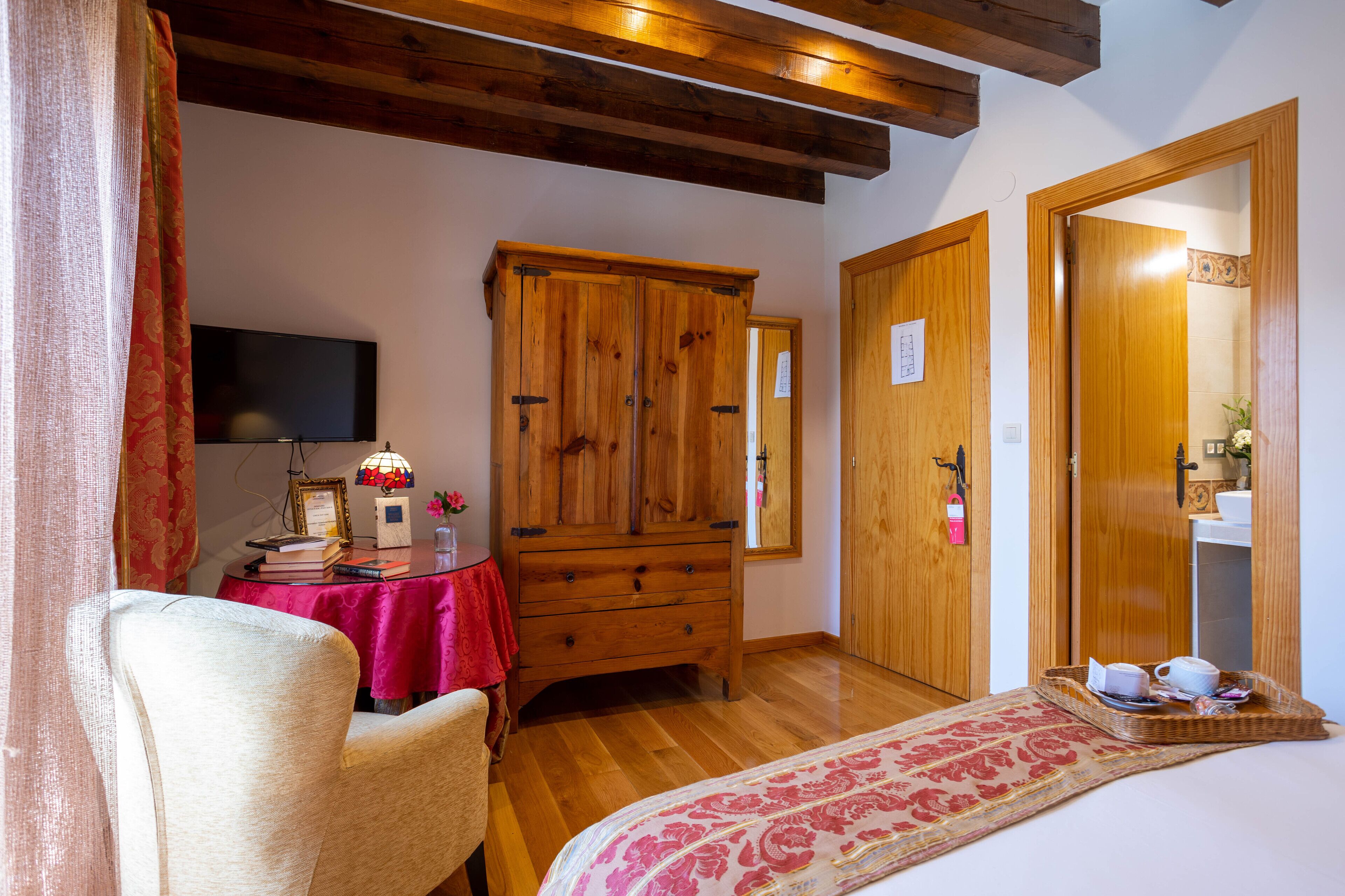 Un dormitorio con un armario de madera, una cama con colcha estampada, una silla, una mesita con mantel rojo y un televisor en la pared.