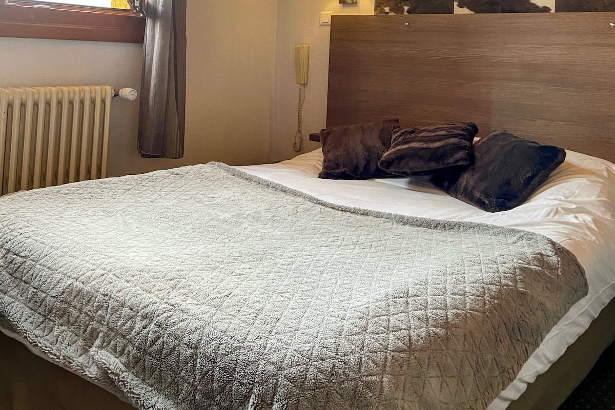 Habitación de hotel con techo de madera, cama con colcha blanca, cabecera estampada con diseño de camuflaje, lámpara de pared y ventana con cortinas.