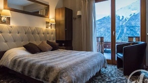 Habitación de hotel con una cama grande, cabecera de madera, dos lámparas de pared, un armario de madera y un balcón con vistas a la montaña.