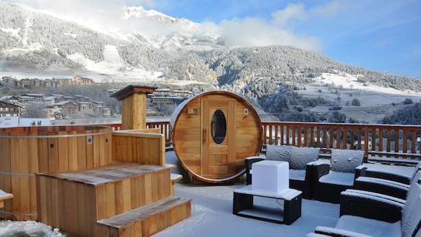 Una sauna de madera con puerta circular, escalones de madera y asientos exteriores con cojines cubiertos de nieve.