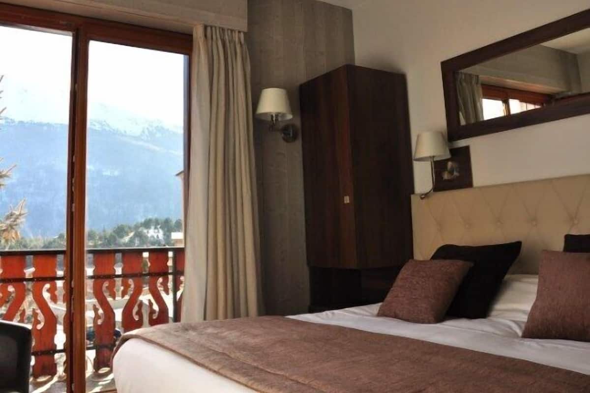 Habitación de hotel con una cama grande, un armario, un balcón con vistas a montañas y dos lámparas fijadas a la pared.