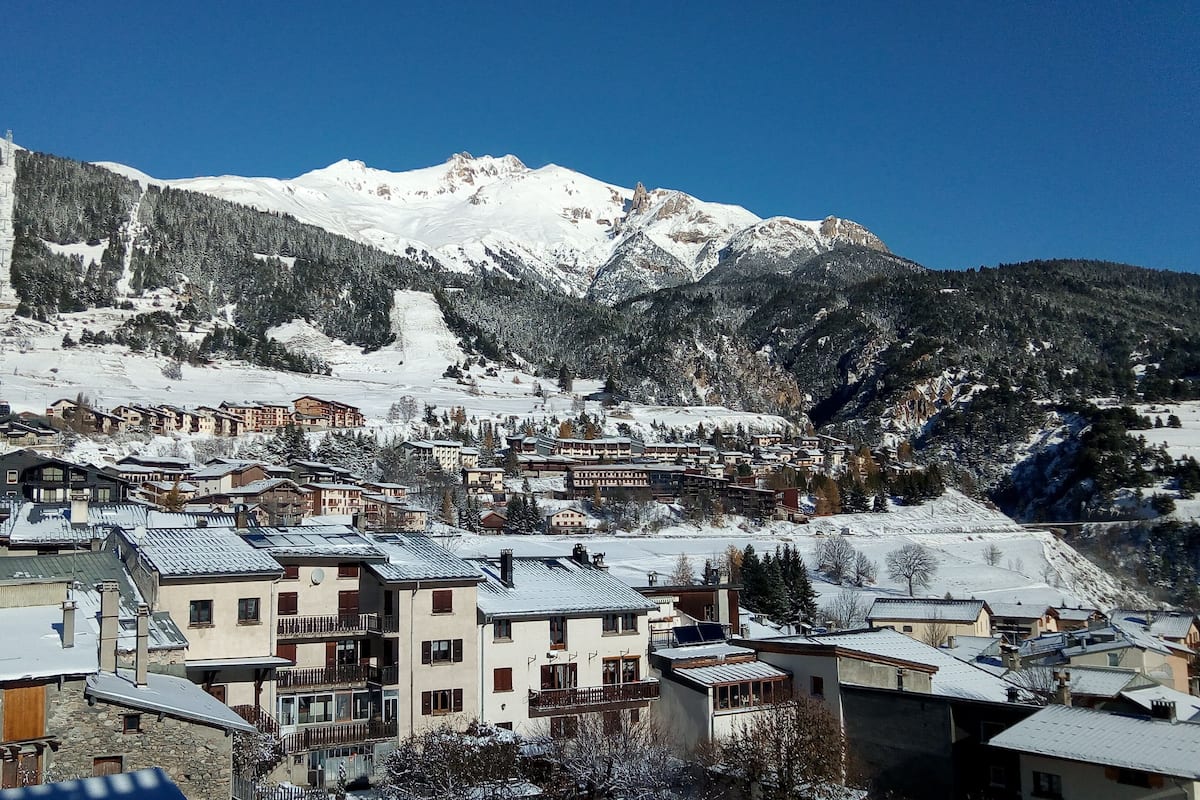 Un pueblo de montaña con edificios nevados y una pista de esquí al fondo.