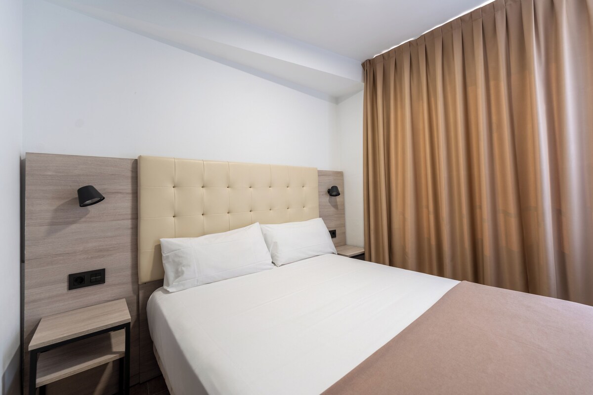 Una cama bien hecha con ropa blanca, un cabecero con diseño acolchado y una mesita de noche con una lámpara.