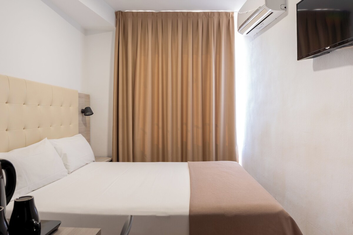 Habitación de hotel con cama, cabecera, mesita de noche con lámpara y televisor montado en la pared.