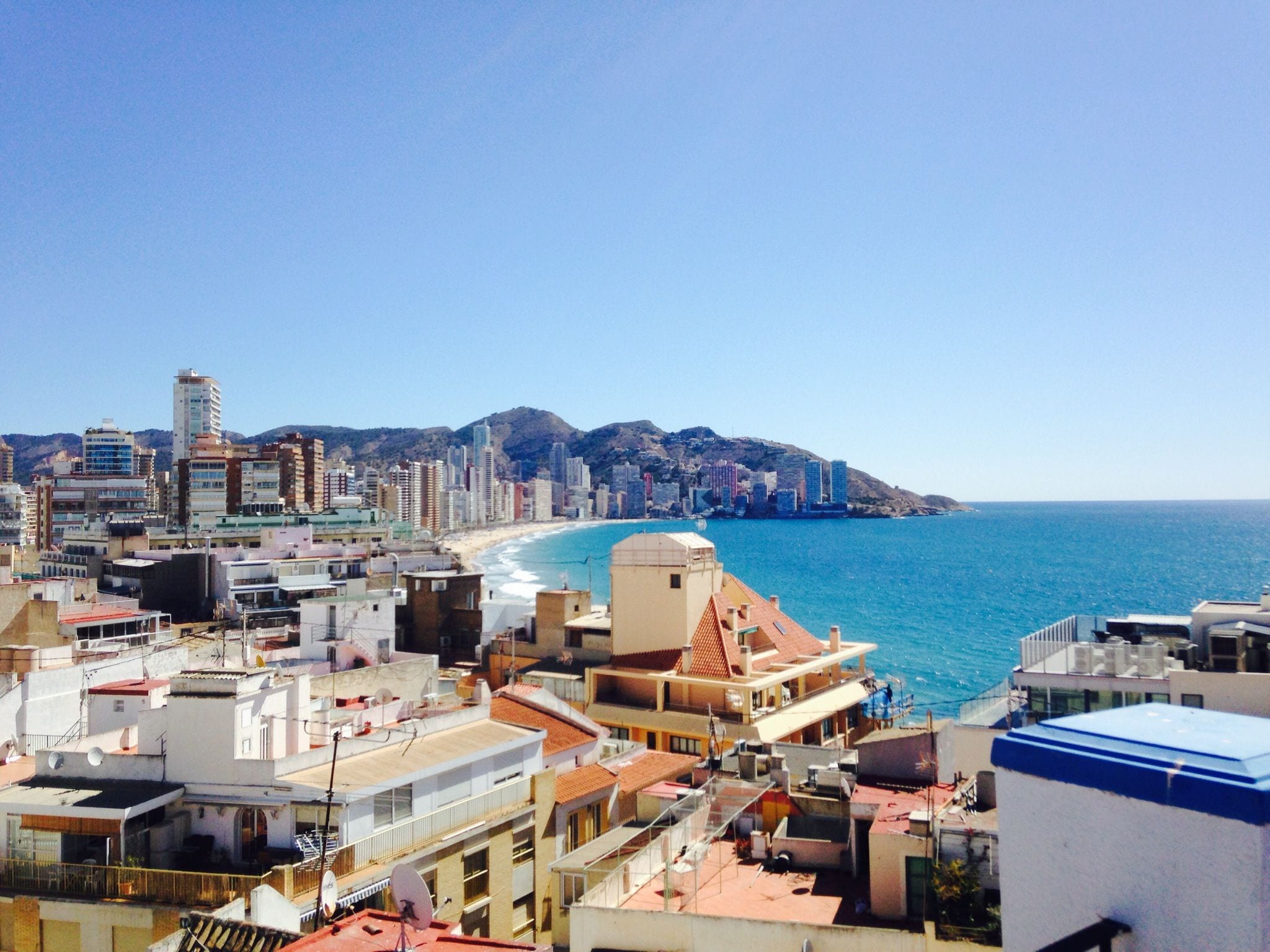A coastal cityscape with a mix of residential and commercial buildings, a clear blue sky, and a beach in the distance.