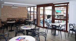 A café with multiple tables and chairs, large windows, and a view of the street outside. - Hotel Queens (Benidorm)
