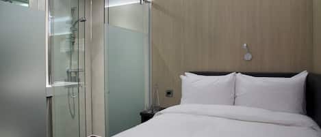 A modern hotel room with a glass-enclosed shower, a bed with white linens, and a wooden accent wall.