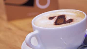 Una taza blanca de café con diseño de latte art sobre un platito, colocada sobre una mesa de madera con un cartel de fondo que dice "CAFE".