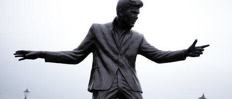 A statue of a man in a suit with outstretched arms, set against a backdrop of a cityscape and a body of water.