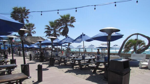 A beachside patio with wooden tables and blue umbrellas, overlooking the ocean.