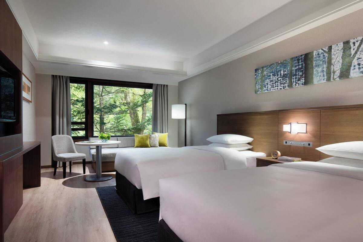 A hotel room with two beds, a desk, a chair, a TV, and a window with a view of trees.