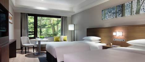 Quarto de hotel com duas camas, uma escrivaninha, uma cadeira, uma televisão e uma janela com vista para árvores.