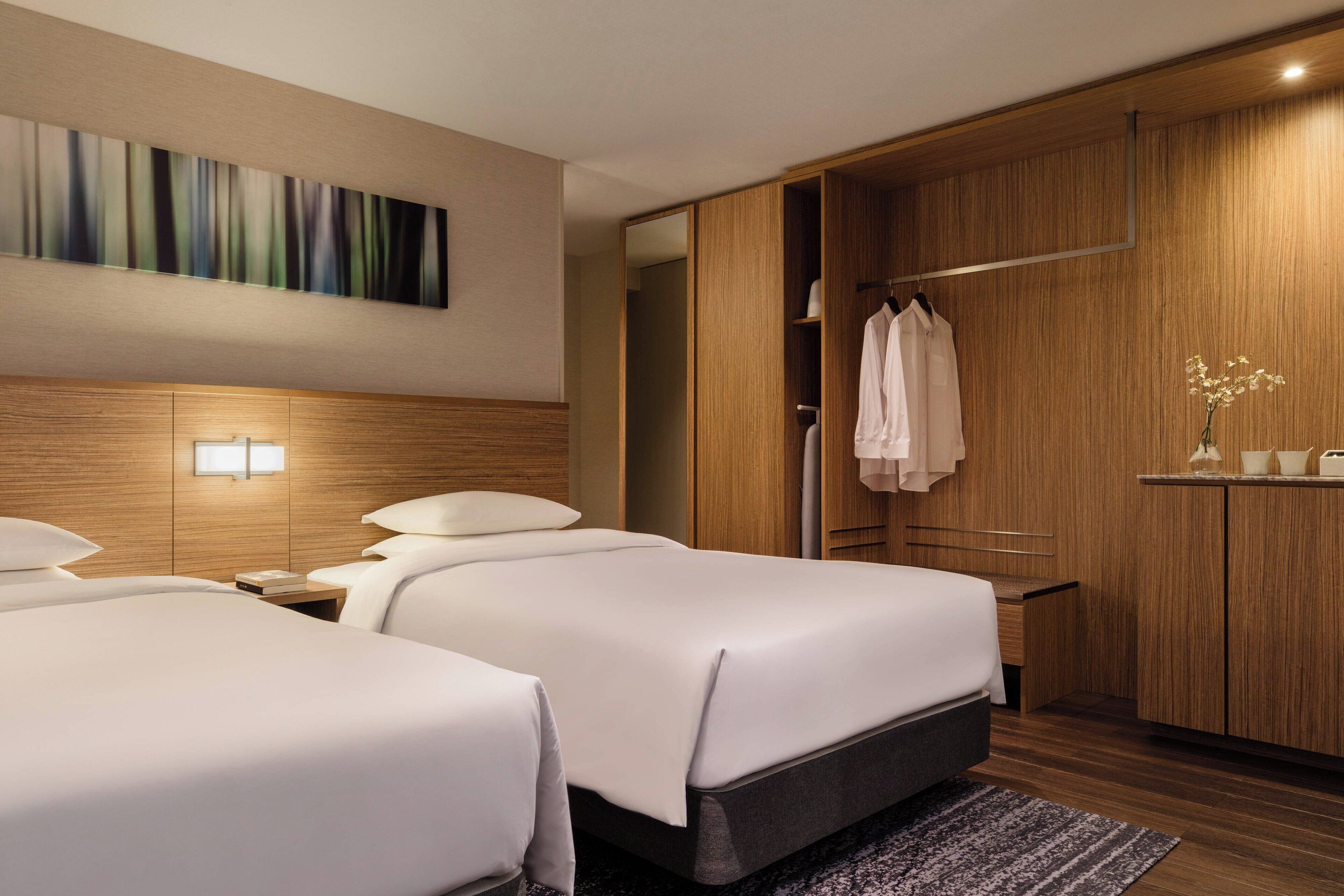 A hotel room with two beds, a wooden wardrobe, a painting on the wall, and a small table with a vase of flowers.