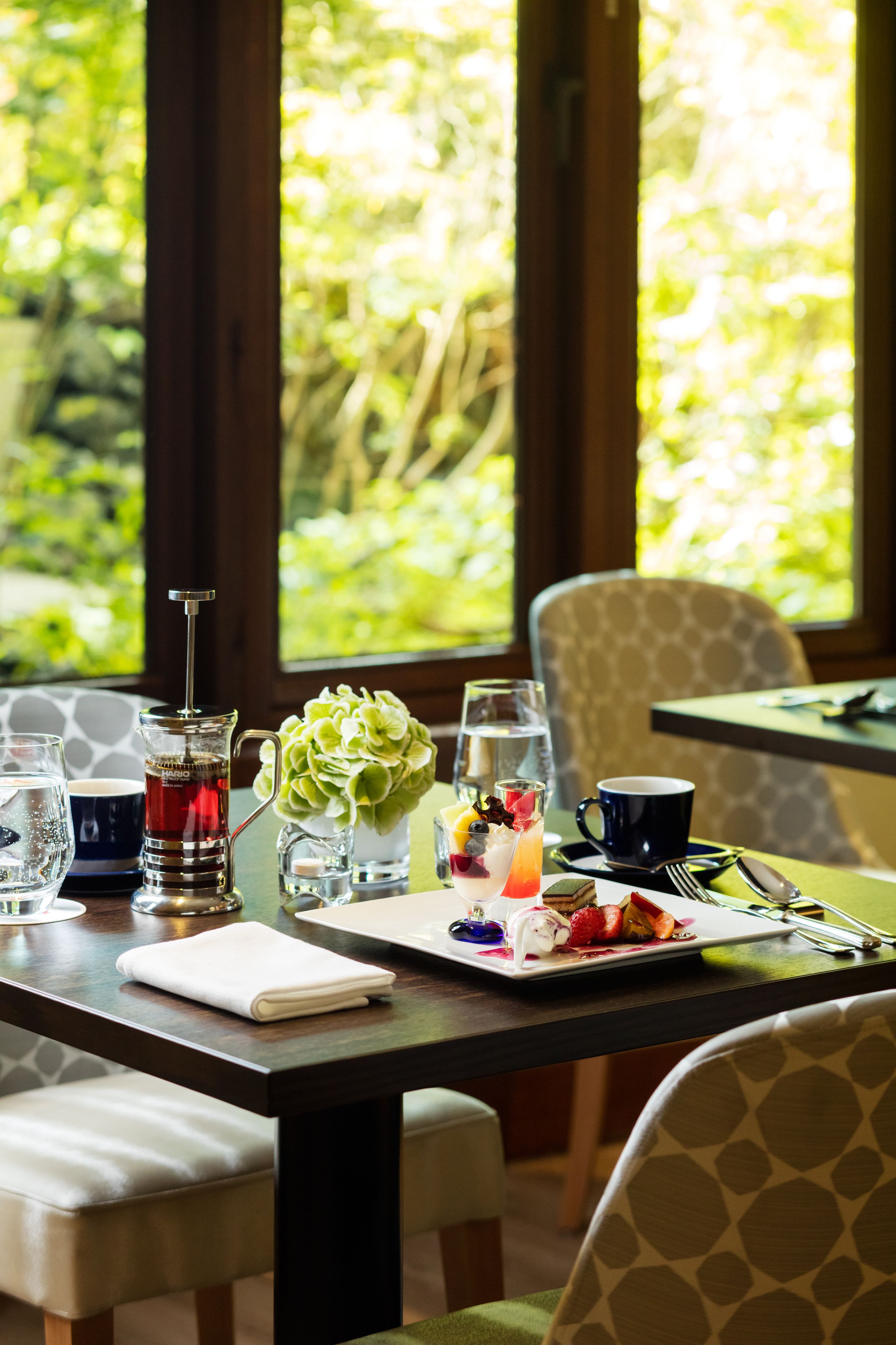 Une table à manger dressée avec une machine à café, un vase de fleurs, un plateau de fruits et une assiette de nourriture.