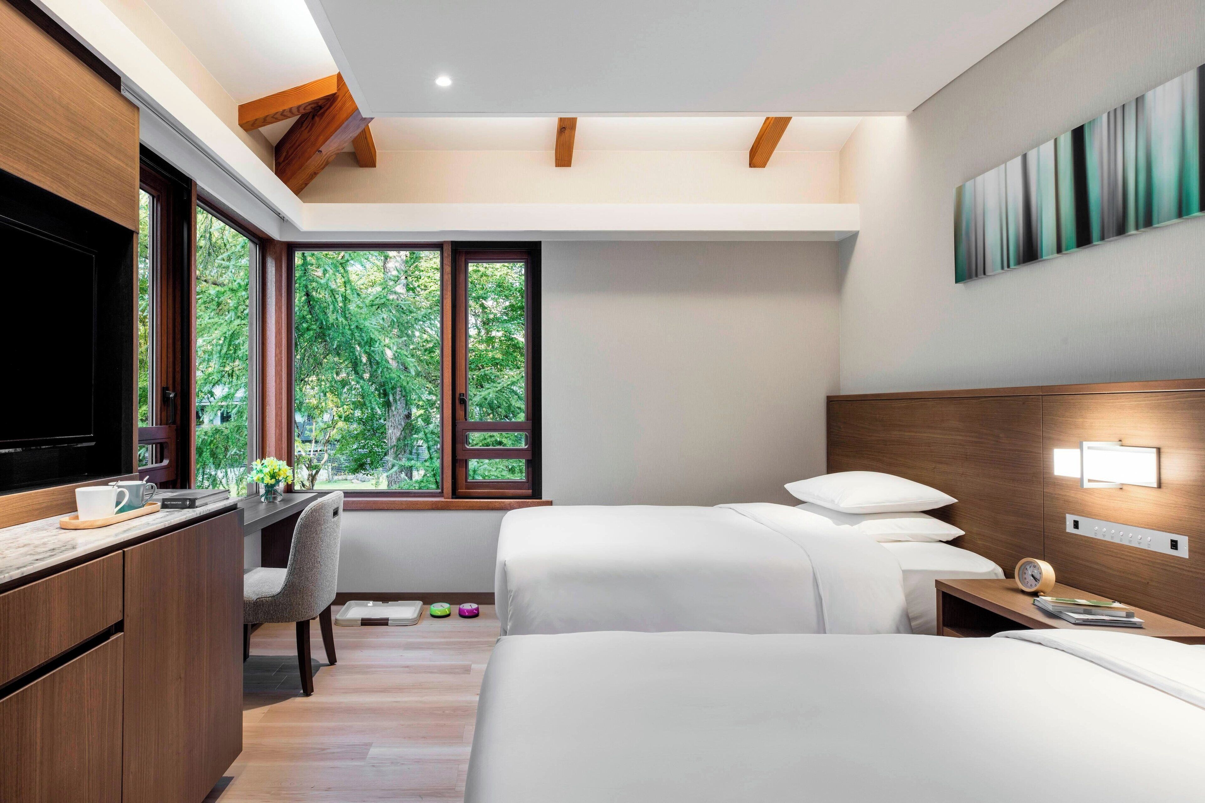 A modern hotel room with two beds, a desk, a TV, and a large window offering a view of greenery.