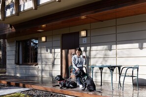 Une personne assise sur une chaise, avec deux chiens tenus en laisse, une table et un bâtiment sur lequel on peut lire l’inscription « A ».