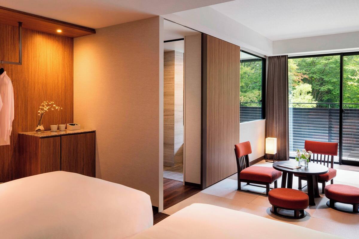 A hotel room with a bed, a seating area with chairs and a table, a wooden cabinet, and a view of trees through large windows.