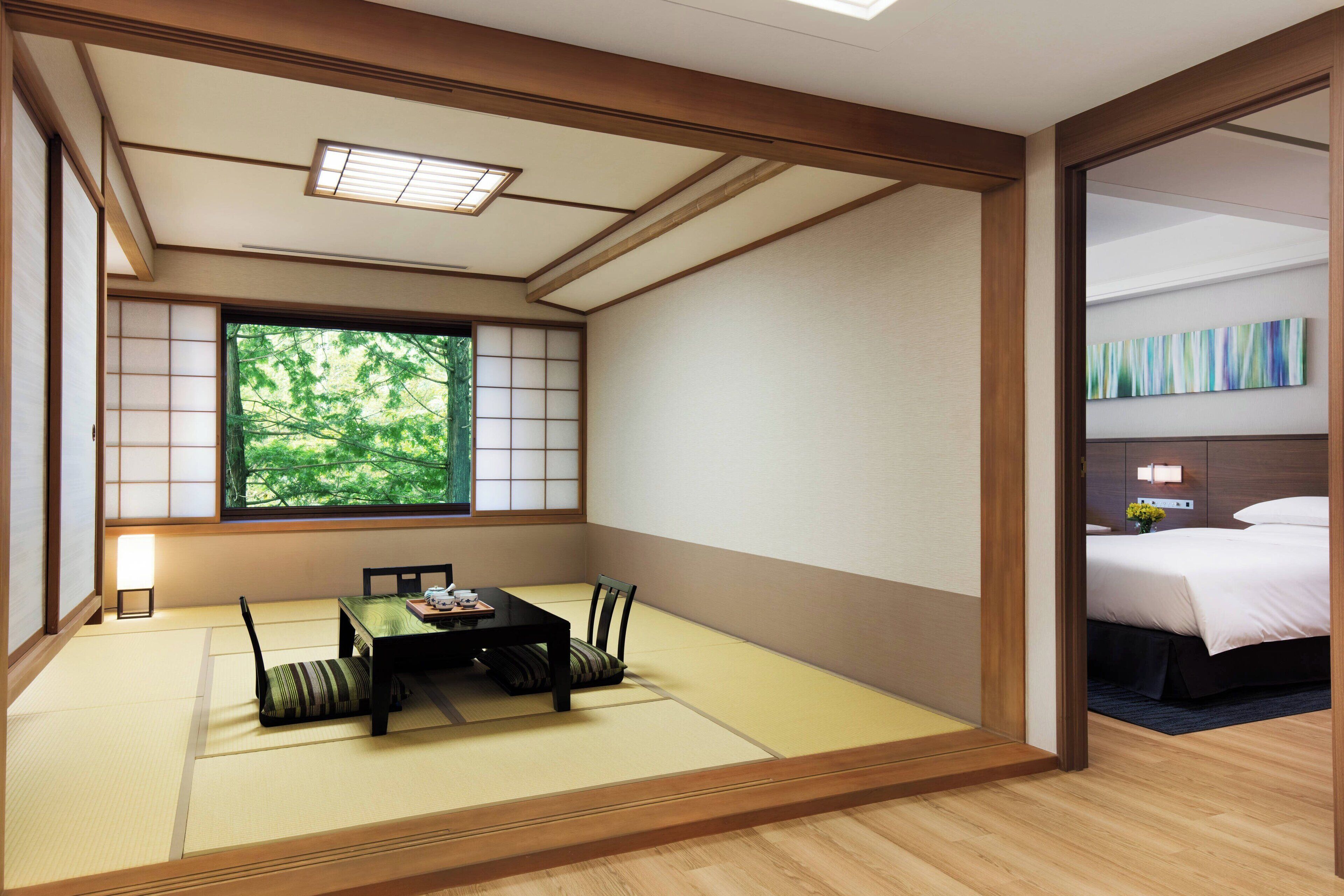 Una habitación japonesa tradicional con suelo de tatami, una mesa baja, sillas, un divisor shoji y una cama con vistas a zonas verdes.