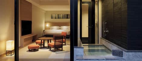 Una habitación de hotel moderna con pared de vidrio, zona de estar con sofá y sillones, cama, televisor y pequeña piscina.