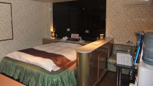 A hotel room with a bed, a desk, a chair, a TV, and an air conditioning unit. - Que Sera Sera - Adults Only (Neyagawa)