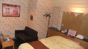 A hotel room with floral wallpaper, a bed with a floral bedspread, a small table with a bowl, a chair, and a mirror with a towel rack. - Que Sera Sera - Adults Only (Neyagawa)