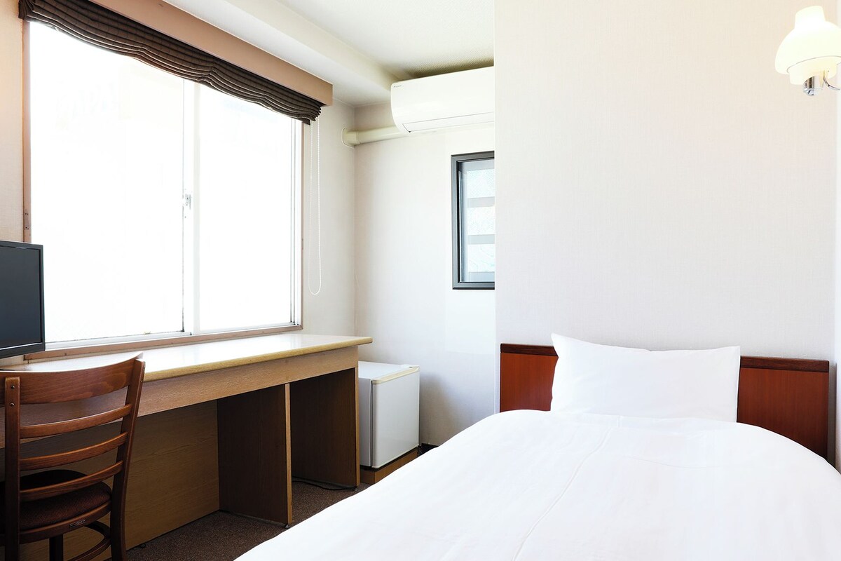 A hotel room with a bed, a desk with a chair, a television, and a window with curtains.