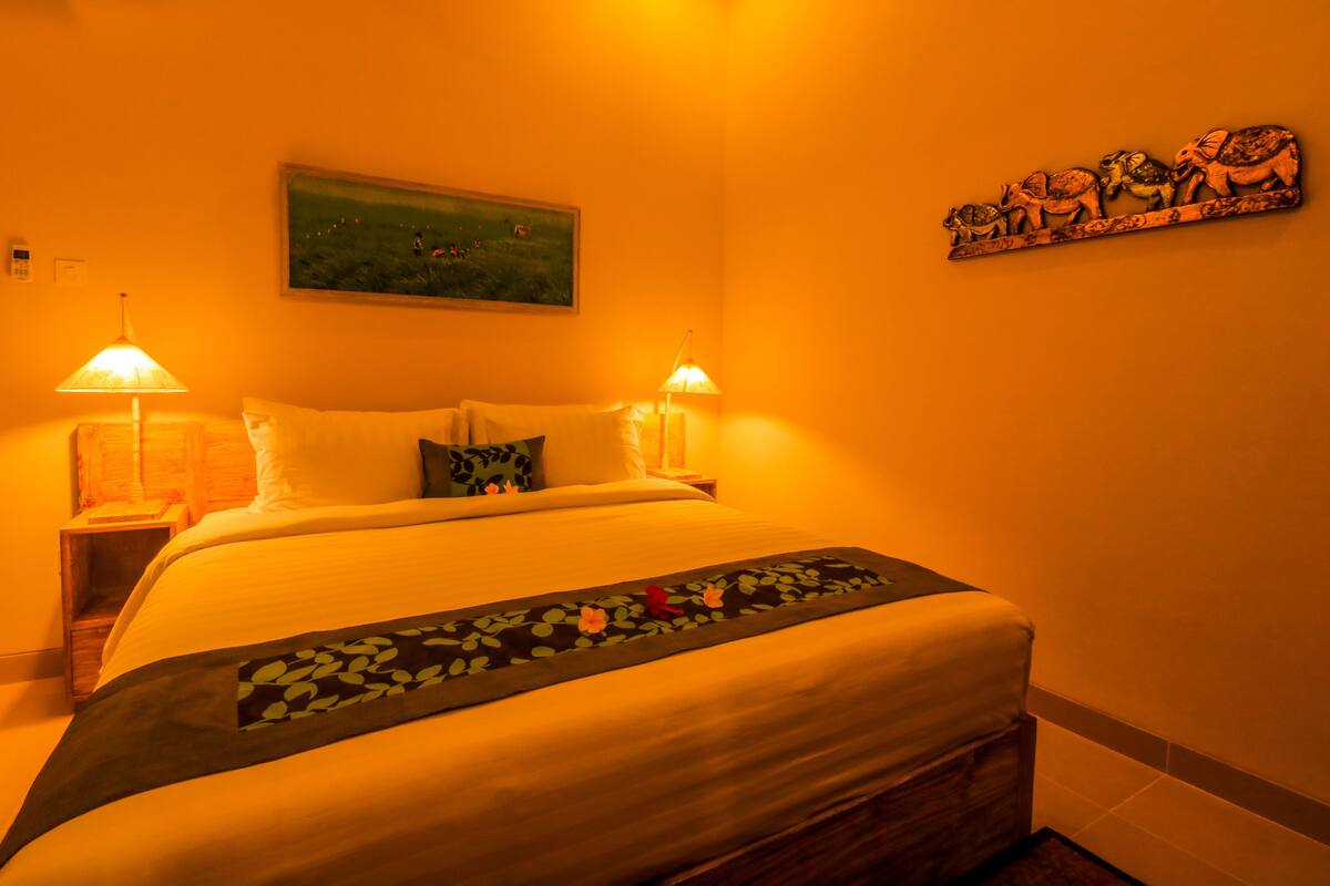 A hotel room with a large bed, two bedside lamps, a painting on the wall, and a decorative piece above the bed.