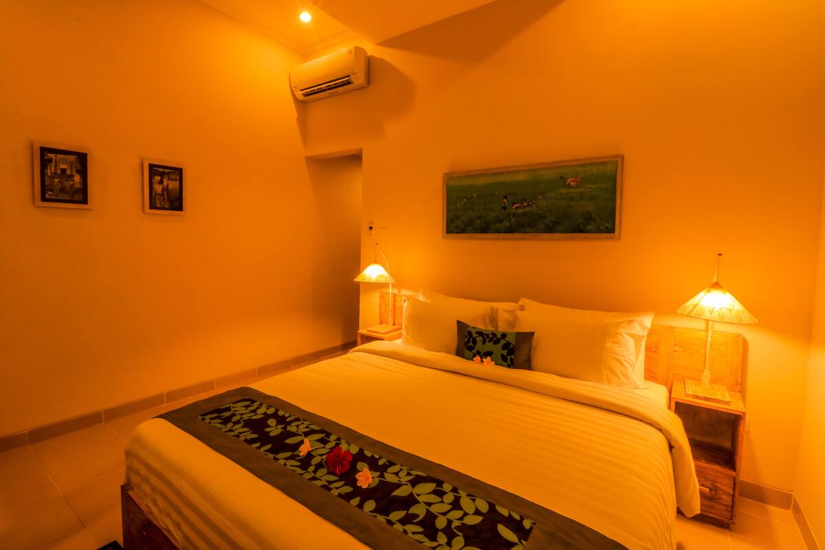 A hotel room with a bed, two bedside lamps, an air conditioner, and two framed pictures on the wall.