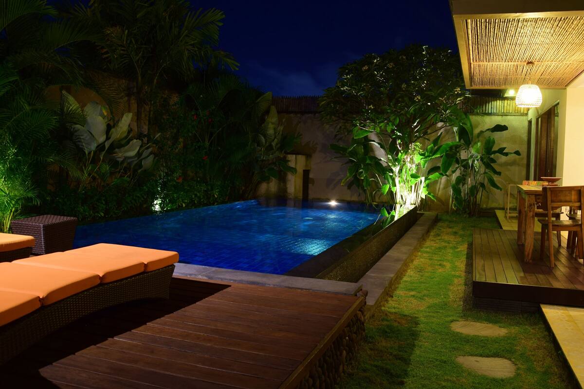 A nighttime swimming pool area with a wooden deck, lounge chairs, and a dining area with a table and chairs.