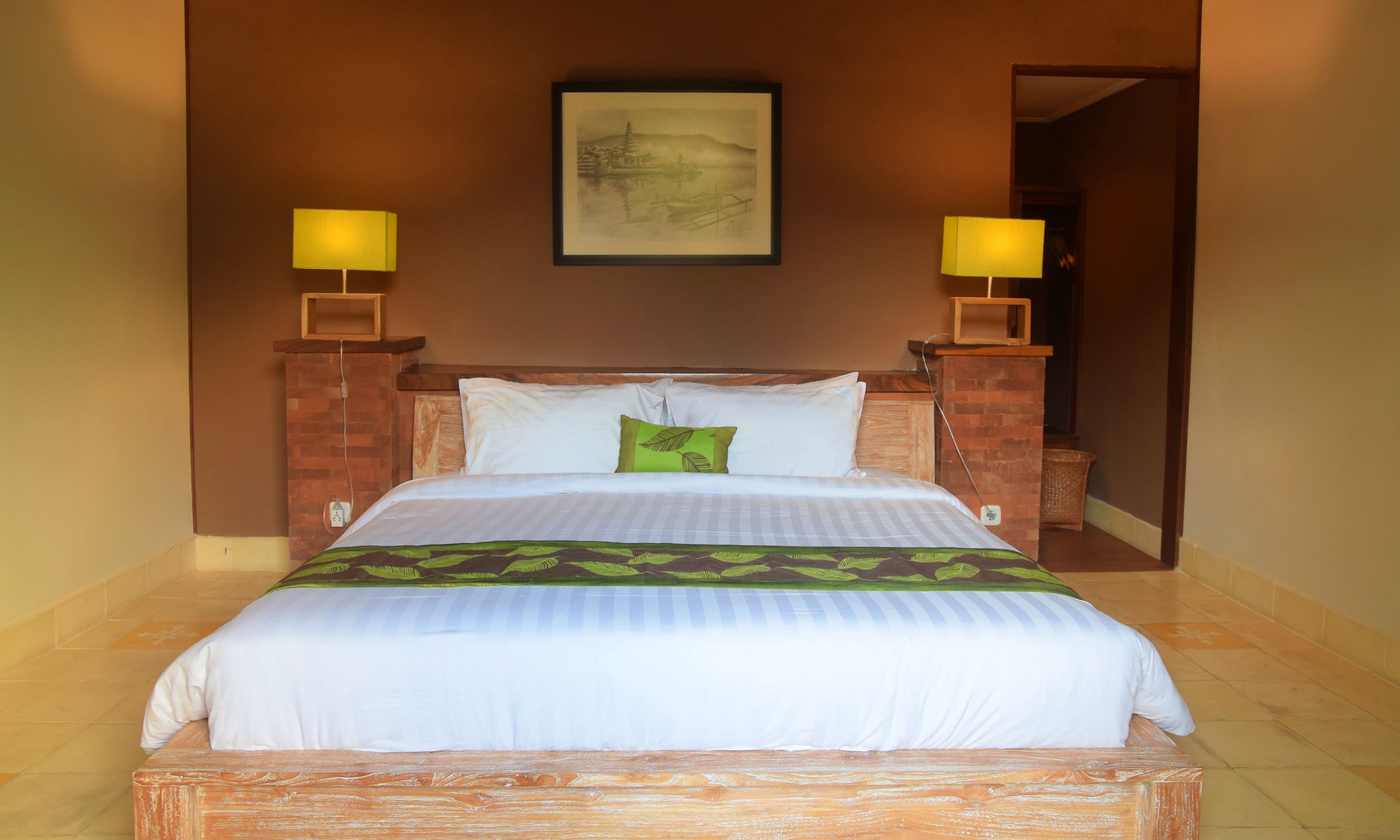 A neatly made bed with white bedding and a green leaf-patterned pillow, flanked by two bedside lamps on brick stands, in a room with a brown wall and a framed artwork above the bed.