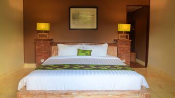 A neatly made bed with white bedding and a green leaf-patterned pillow, flanked by two bedside lamps on brick stands, in a room with a brown wall and a framed artwork above the bed.
