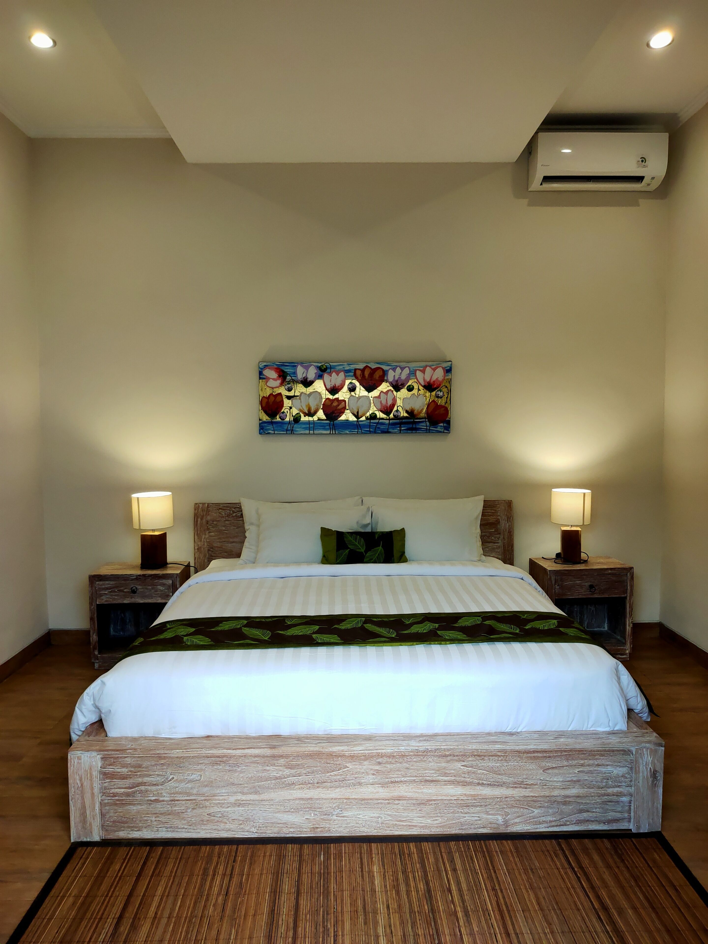 Cama bem arrumada com lençóis brancos, duas mesas de cabeceira com abajures, ar condicionado fixado na parede e um quadro colorido sobre a cama.