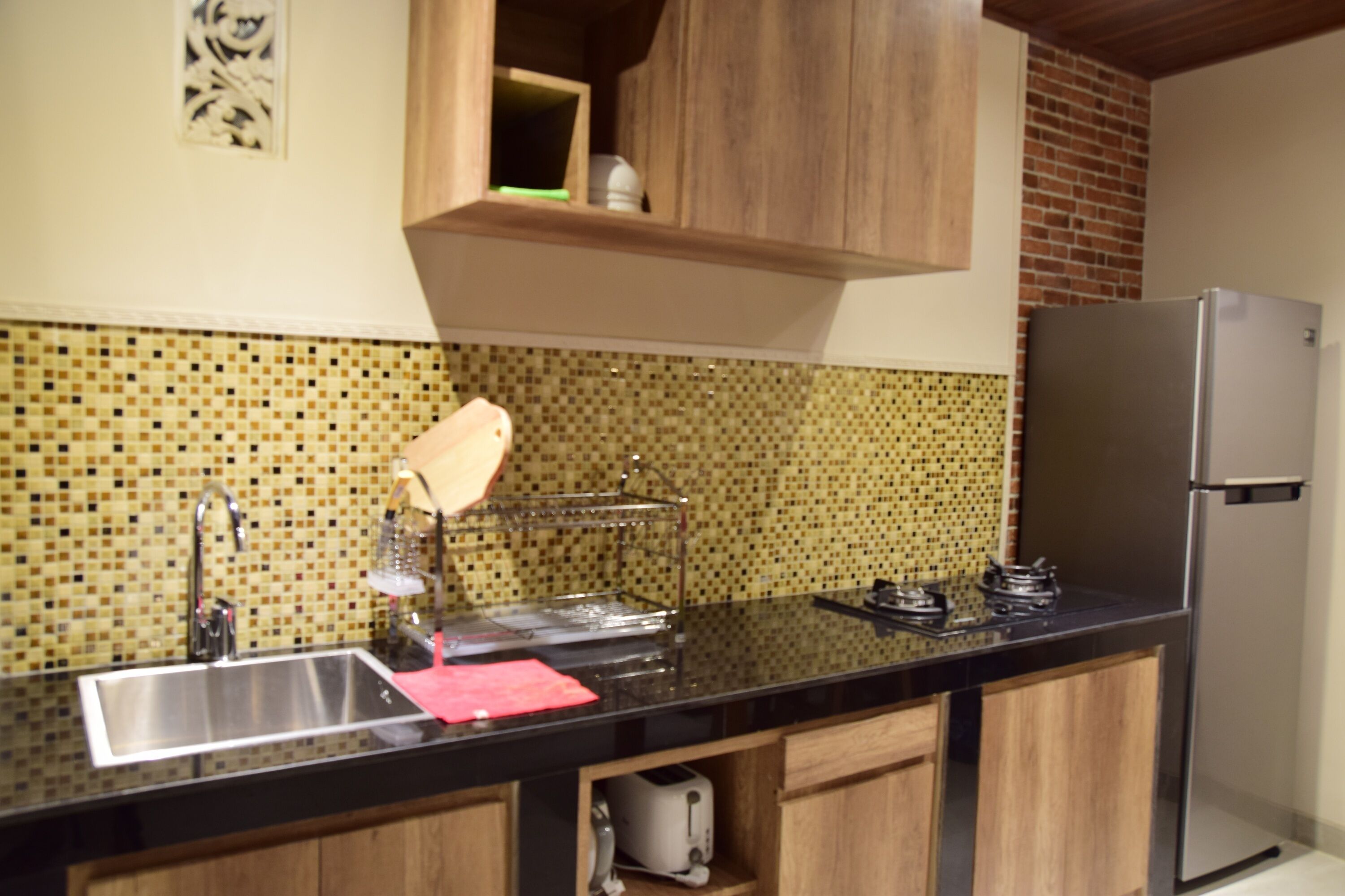 Una cocina con un lavamanos de acero inoxidable, encimera negra, armarios de madera, refrigerador de acero inoxidable y salpicadero de mosaicos.