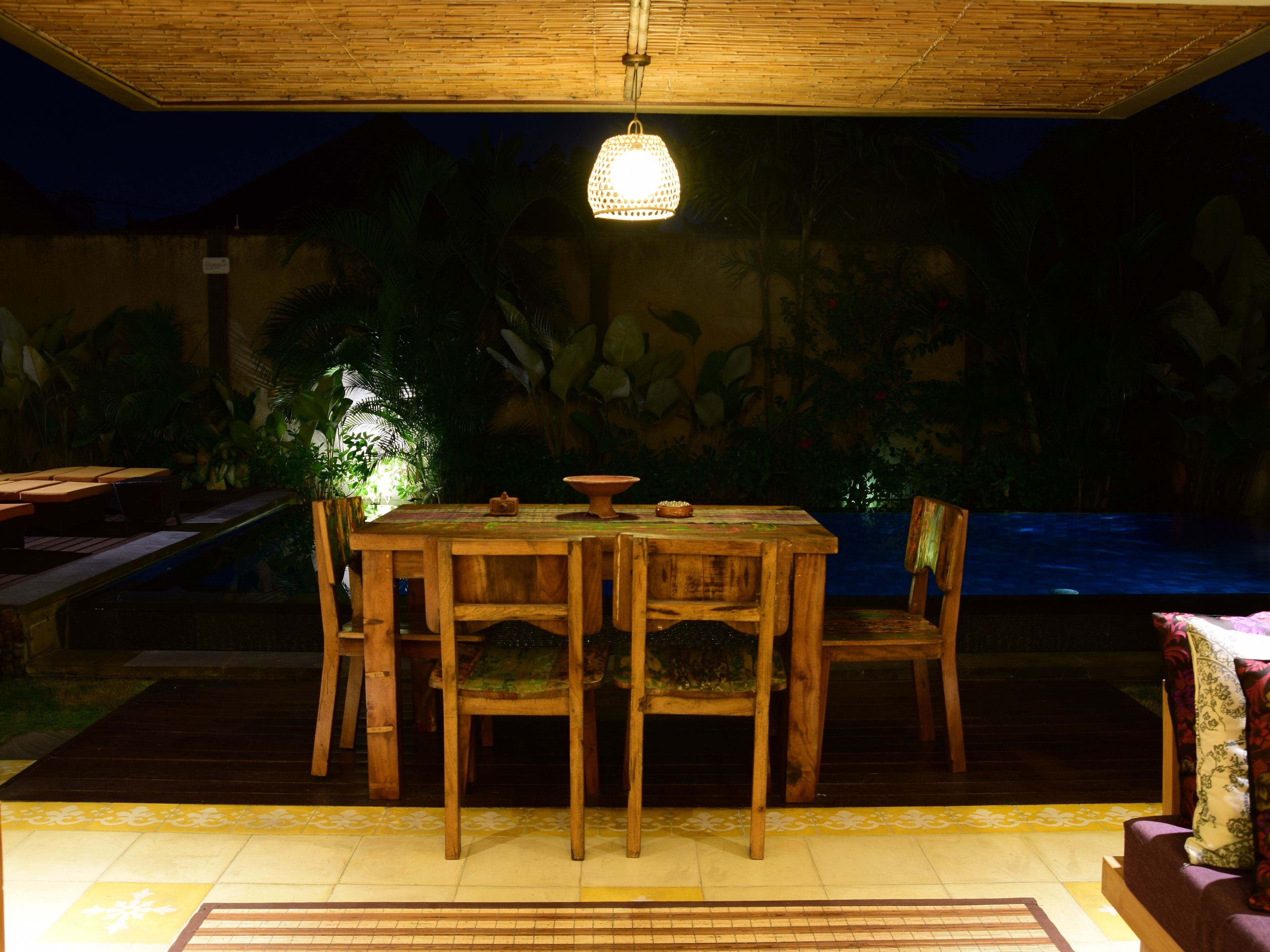 Un área de comedor al aire libre con mesa y sillas de madera, un colgante de luz y vista a una piscina.