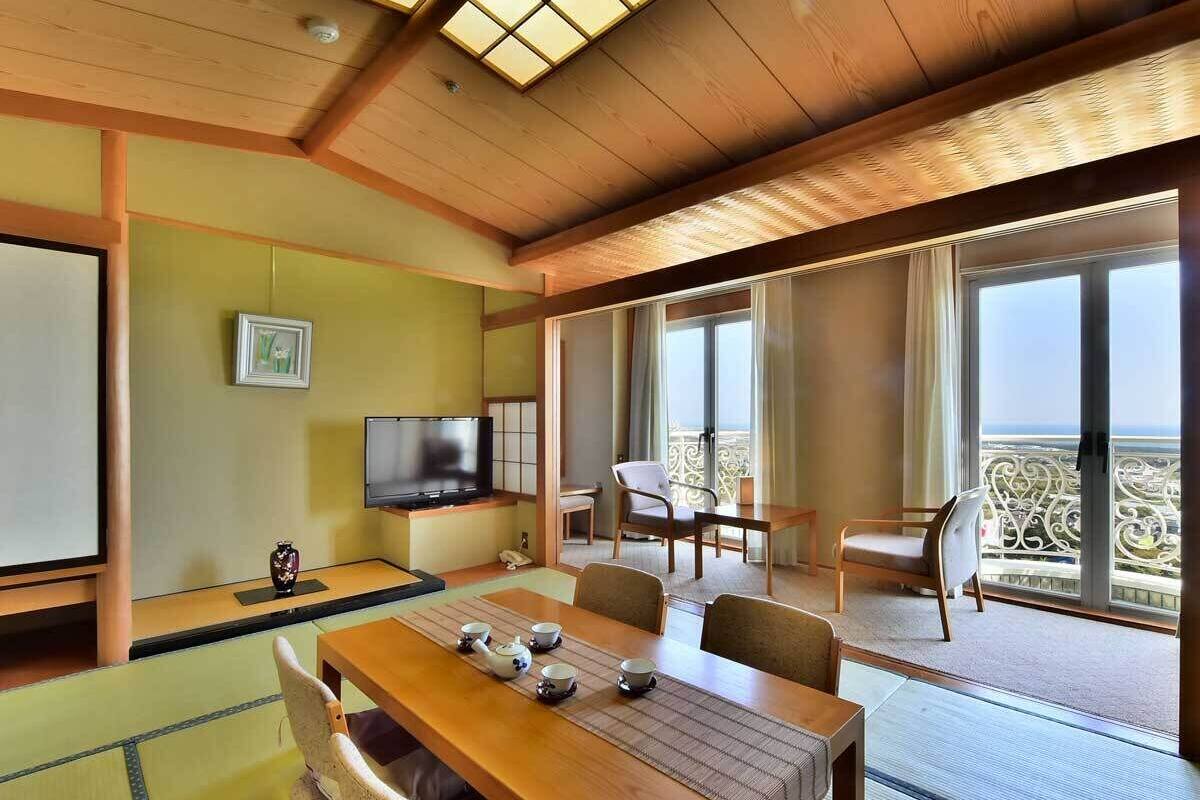 Una habitación tradicional japonesa con suelo de tatami, una mesa baja con juego de té, un televisor y un balcón con vistas.