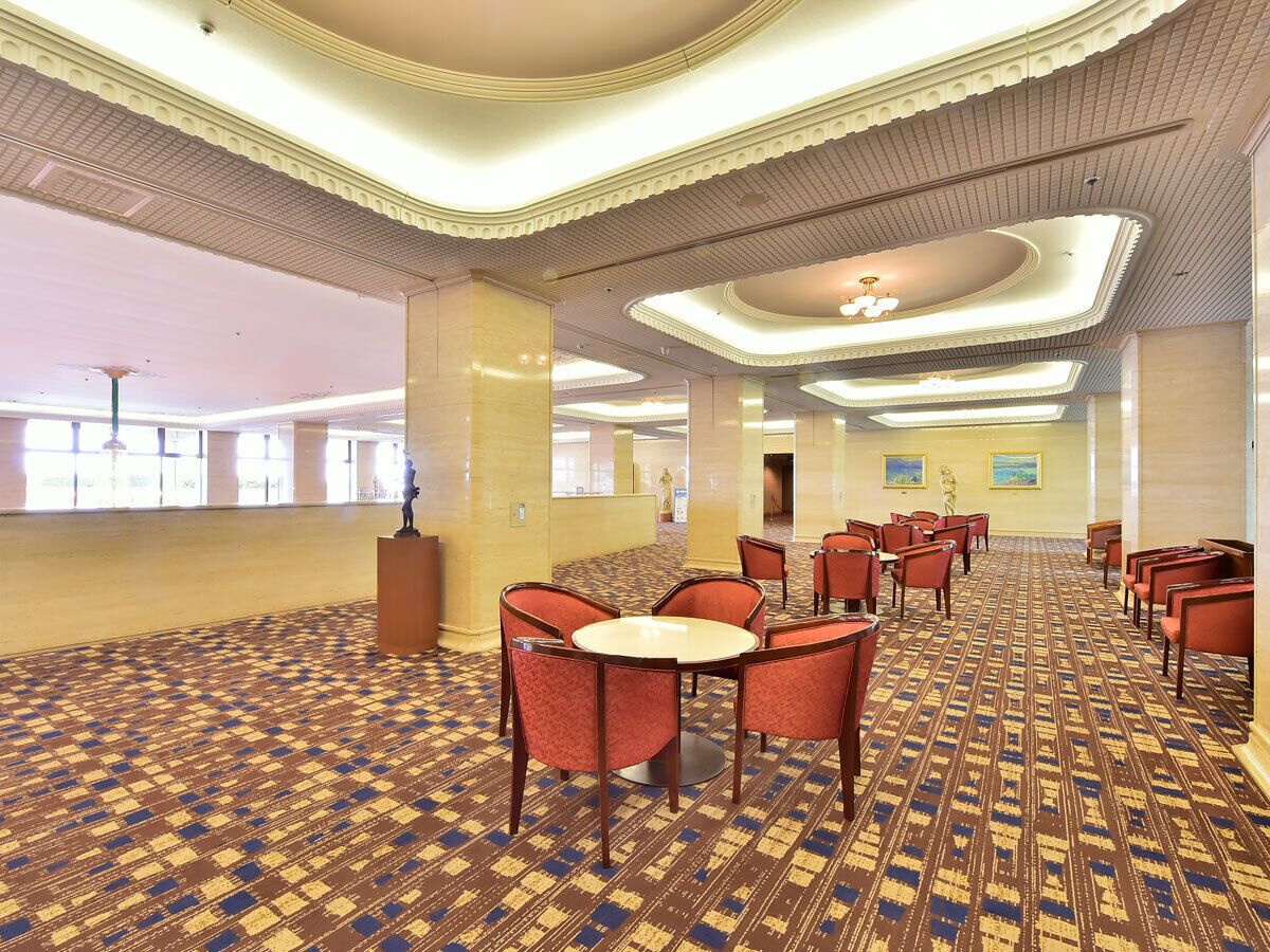 Um saguão de hotel com carpete estampado, mesas redondas com cadeiras e teto alto com molduras decorativas.