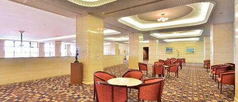 A hotel lobby with a patterned carpet, round tables with chairs, and a high ceiling with decorative moldings.
