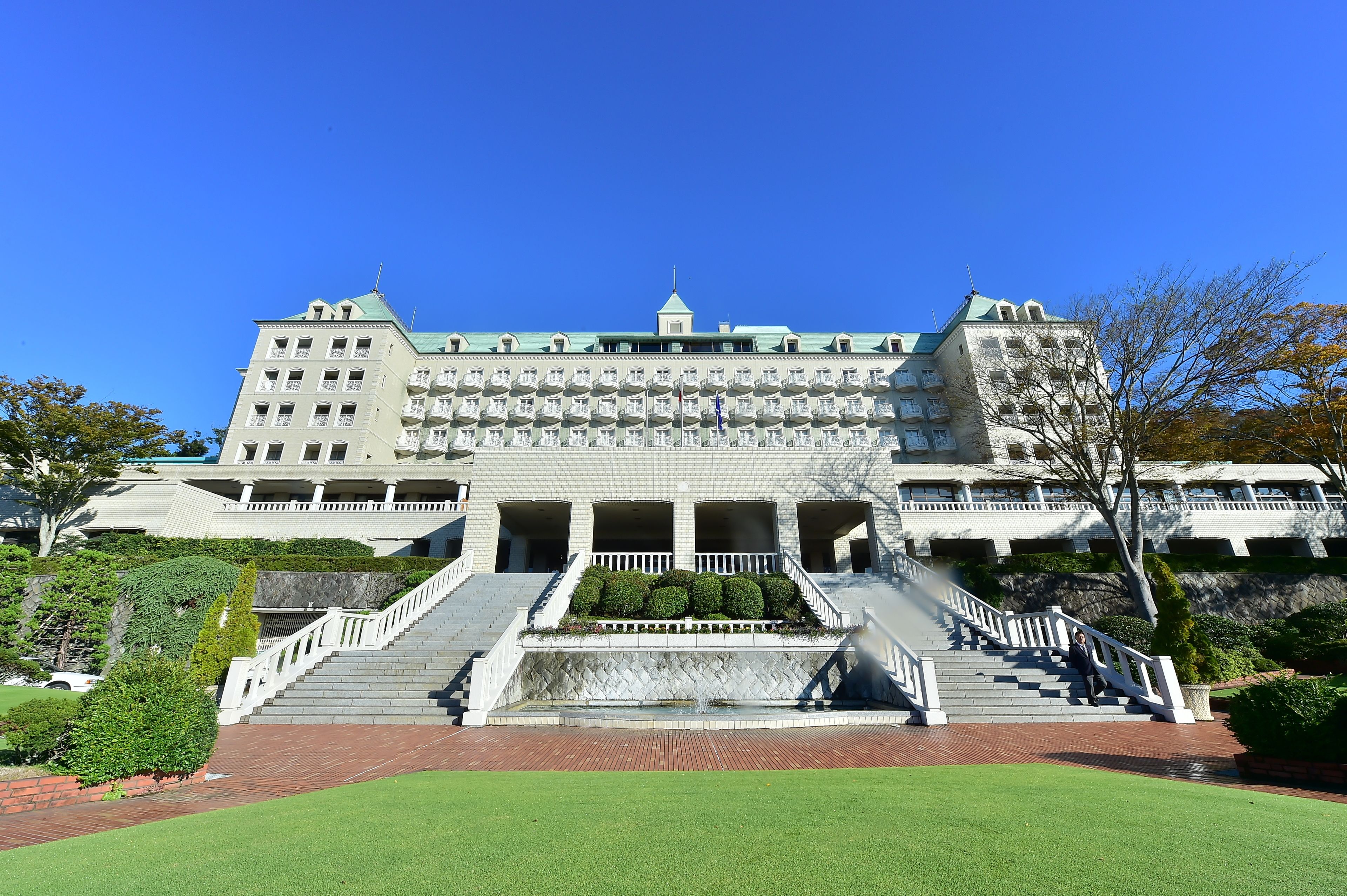 A large, multi-story building with a central staircase leading to a balcony, surrounded by well-maintained lawns and a small fountain.