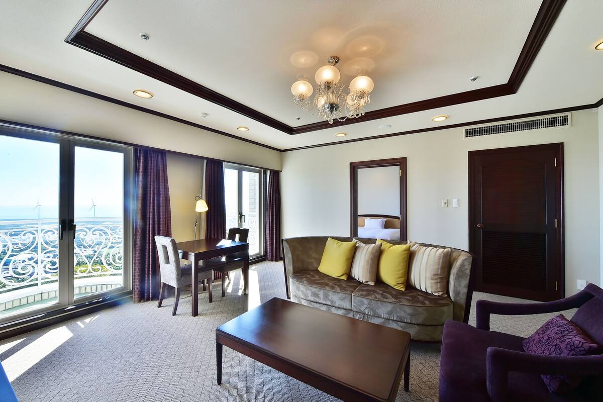 Habitación de hotel con balcón amplio, sofá, sillón, mesa y mesita de centro.
