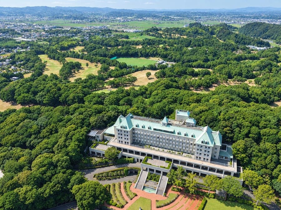 A large, multi-story building with a symmetrical facade, surrounded by extensive greenery and a well-maintained lawn.