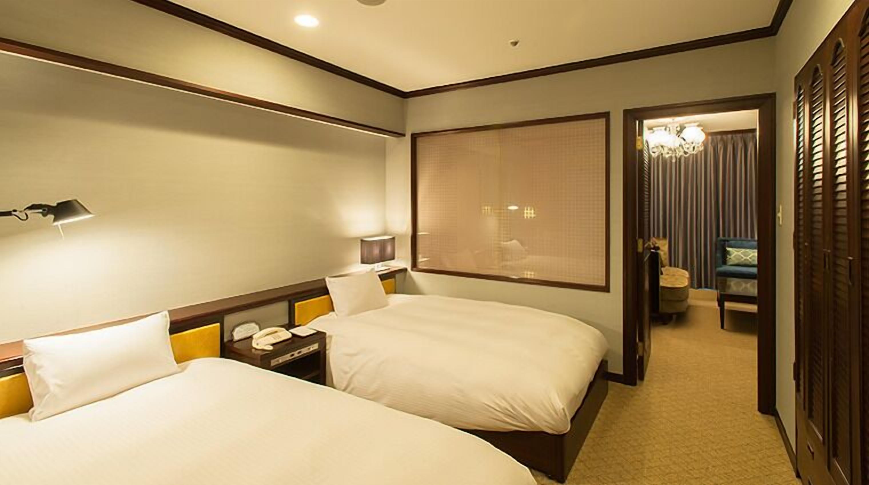 A hotel room with two beds, a bedside table with a lamp, a telephone, and a window with blinds.